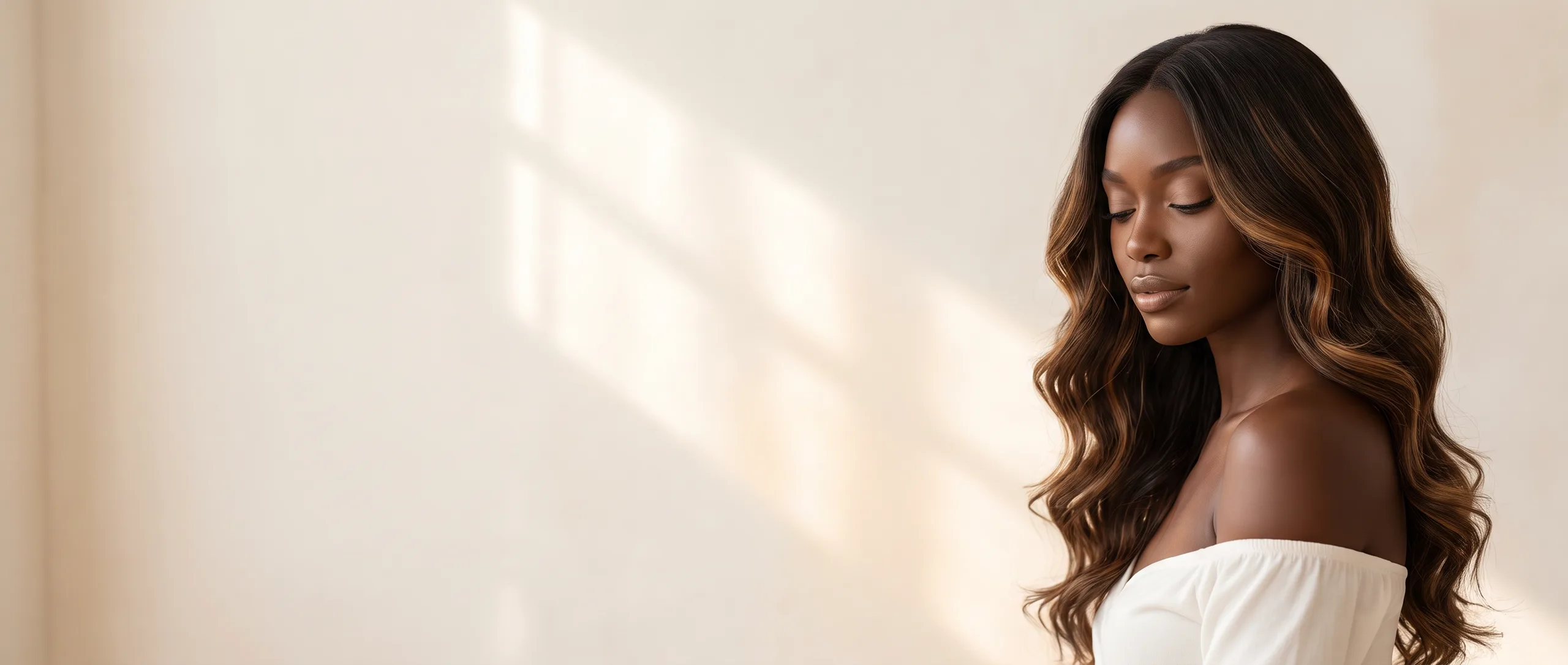 Model with long caramel balayage waves against a sunlit cream wall